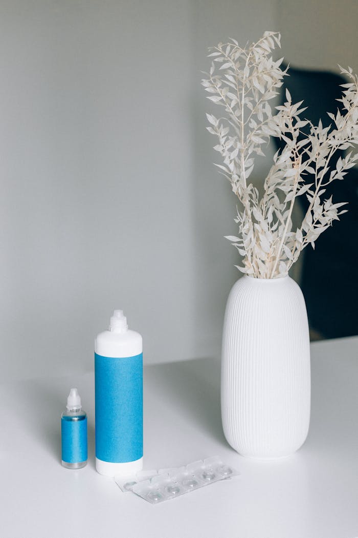 Close-up of eye care bottles and a white vase with dried leaves on a table.